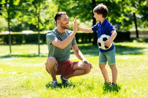 A father and his young son high-five on a green lawn outdoors; the boy holds a soccer ball. The scene shows warm parental encouragement after a successful game and positive bonding from physical activity.