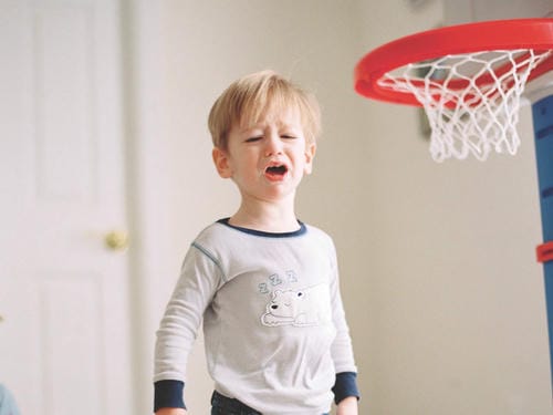 A toddler cries loudly beside an indoor mini basketball hoop, upset from failing to make a shot and overcome by frustration.