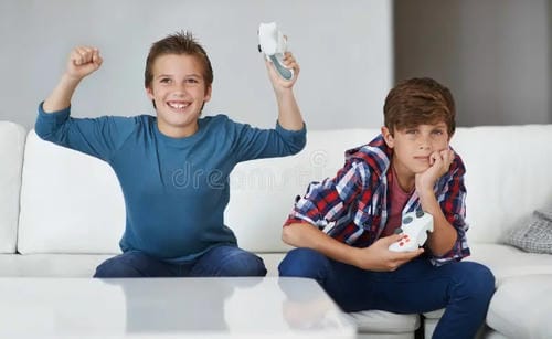 Two boys play video games on a sofa. The boy on the left cheers excitedly with raised arms after winning, while the other boy looks glum and dejected from defeat, showing opposite moods caused by game victory and loss.