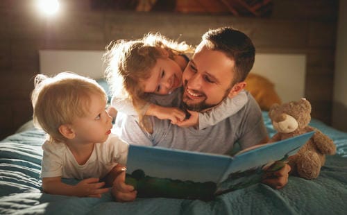 A dad reads a picture book to his two kids on the bed at night; one little girl hugs his neck playfully while the boy listens attentively, sharing cozy family bonding before bedtime.