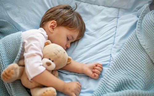 A small child curls up in bed, hugging a stuffed teddy bear and sleeping soundly under a soft knit blanket, enjoying a restful night’s sleep.