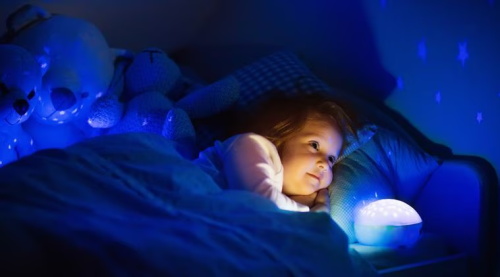 A little girl lies in bed at night, gazing gently at a star-projection nightlight that casts star shapes across the dark bedroom wall, with plush teddy bears beside her pillow, feeling peaceful before sleep.