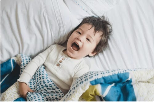 A young toddler stretches and yawns happily on white bed linen, full of good mood after waking up from a night’s rest.