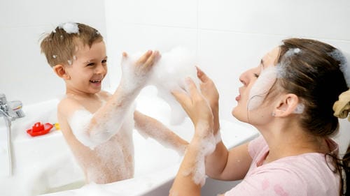 A mother and her little boy play happily with fluffy bath foam in the bathtub, tossing soap bubbles at each other during a joyful evening bath routine.