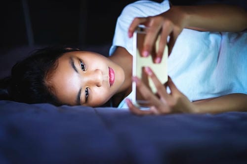A girl lies on bed at night, glued to her cellphone screen, staying up late scrolling on mobile in dim surroundings.