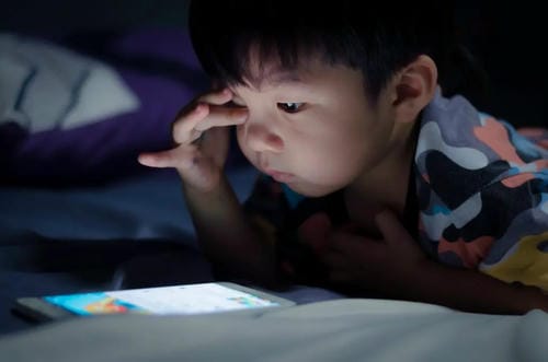 A young boy lies on bed in total darkness, focusing intently on a glowing tablet screen; the bright light strains his eyes, a typical case of excessive screen use before bedtime.