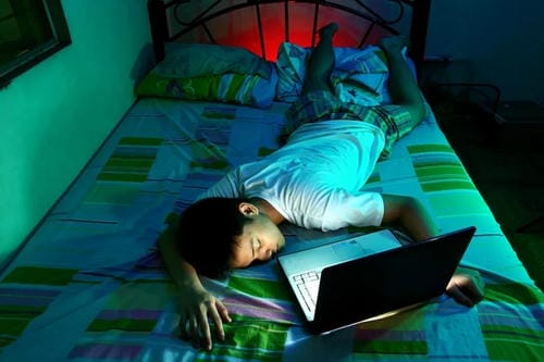 A boy passes out face-down on his bed next to an open laptop after staying up late with electronic devices, worn out from prolonged screen time.
