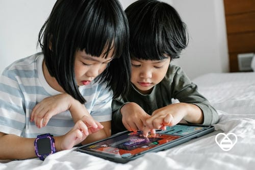 Two little Asian girls lie side by side on a bed, tapping a tablet screen together and focusing on interactive digital games, sharing screen time at home. One girl wears a kids’ smartwatch on her wrist.