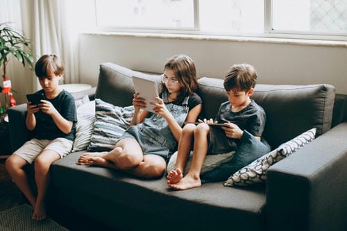 Two boys and one girl sit together on a home sofa; each child focuses separately on their own smartphone or tablet, immersed in personal screen activities instead of talking face to face.