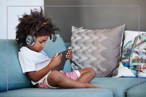 A young boy sits comfortably on a living-room couch with over-ear headphones on, fully absorbed in content on his tablet while relaxing indoors.