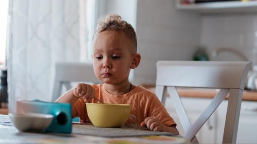 A small toddler sits at a dining chair, holding a spoon and feeding himself from a yellow bowl with no electronic devices nearby, forming a good eating habit.