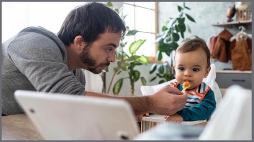 A father feeds his infant with a spoon, while a tablet is placed on the table in the foreground; the baby focuses on the screen instead of the food being offered.