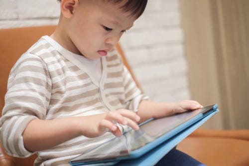 A little boy sits by the sofa, fully concentrated on tapping a tablet screen, immersed in solo digital entertainment.