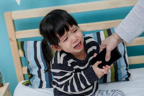 A little girl sits on bed crying and clinging tightly to a smartphone, screaming as an adult tries to take the device away, having formed strong electronic device dependence.
