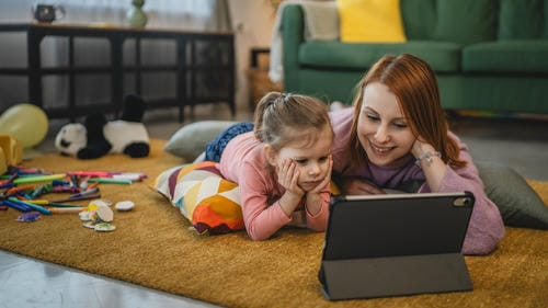 A mother and her little girl lie side by side on the living room carpet, leaning their heads to view content on a shared tablet amid scattered art supplies.