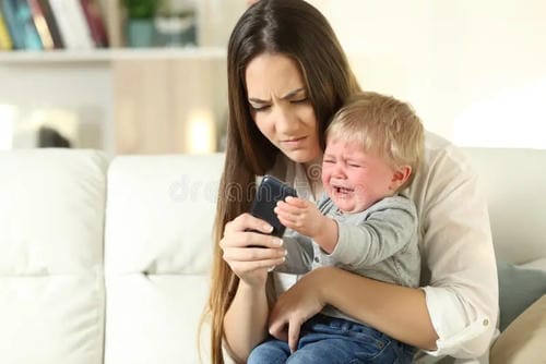 Held in mother’s arms, the toddler bursts into tears and reaches desperately for the mobile phone in mom’s hand, throwing a fit to get screen time.