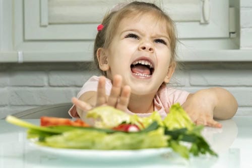 A little girl makes a fierce rejecting gesture with her hand and opens her mouth wide to protest, refusing the plate of fresh vegetable salad.