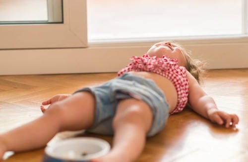 The child lies sprawled on the wooden floor screaming and throwing a fit because she does not want to have her meal.