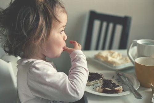 A toddler sits at the dining table and eats bread spread with sweet chocolate crumble by herself, happily enjoying her preferred sweet food.
