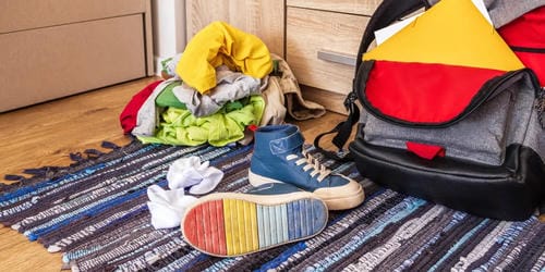 Piled wrinkled clothes, loose socks, casual sneakers and an open duffel bag lie scattered across a woven rug, showing disorganized daily belongings left out on the floor.