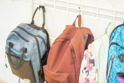 Multiple colorful school backpacks hang tidily from a white grid wall rack, off the floor and neatly stored to save floor space.