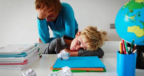 A boy lays his head down on the desk amid piles of exercise books and crumpled waste paper, completely worn out from heavy homework, while an adult beside him also looks worn out.