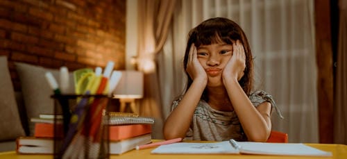 A young girl rests her cheeks in both hands at a desk stacked with textbooks and stationery, looking weary and unmotivated to finish her school assignments.