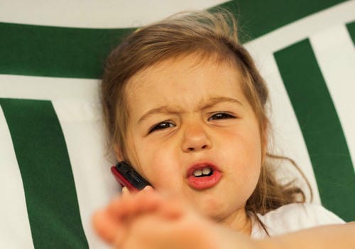 A young girl lies back against a green-and-white striped cushion, holding a toy cellphone to her ear with an annoyed, grumpy facial expression, seemingly throwing a tantrum during pretend phone play.