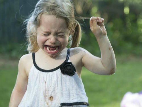 A little blonde girl cries bitterly outdoors, tugging at her hair with one hand. Her dress is stained with mud, suggesting she got upset after falling or having an unhappy accident during outdoor play.
