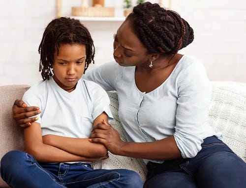 A mother sits on the sofa, gently holding her child’s arms to soothe the upset little girl who crosses her arms tightly and looks sullen after getting into a bad mood.