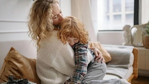 The mother wraps her arms around her tearful little boy on the couch, giving a tight comforting embrace to calm the upset child’s negative emotions.