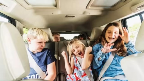 Several children sit in the rear vehicle seats with fastened safety belts; two girls cheer and wave excitedly while a boy beside them looks amused, enjoying a joyful family road trip.