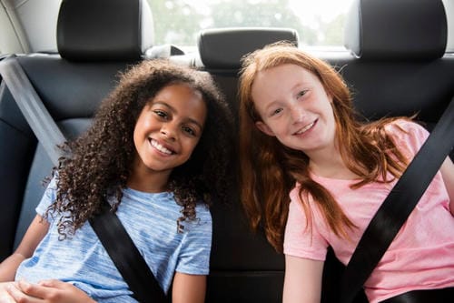 A pair of preteen girls sit side by side on the auto backseat, both wearing standard safety belts and smiling happily for the camera.