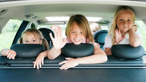 Three young girls lean their heads over the backseat headrests and greet outward with friendly waves, looking cheerful during the car ride.