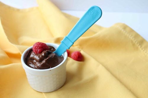 A small ceramic ramekin holds thick chocolate mousse topped with fresh raspberry, placed on warm yellow fabric as a sweet kids’ dessert option.
