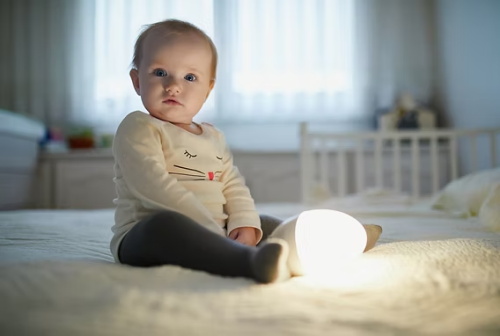 A baby sits alone on a bed beside a glowing night lamp, wide awake instead of sleeping, illustrating a common toddler bedtime wakefulness issue.