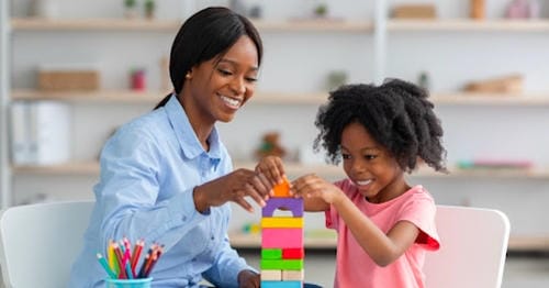 An educator/mom and a little girl cooperate to stack bright-colored wooden building blocks; both beam happily during the focused hands-on educational play.