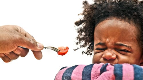 A little kid screws up their face and shuts eyes tightly, resisting a fork with tomato held by an adult and rejecting this vegetable food.