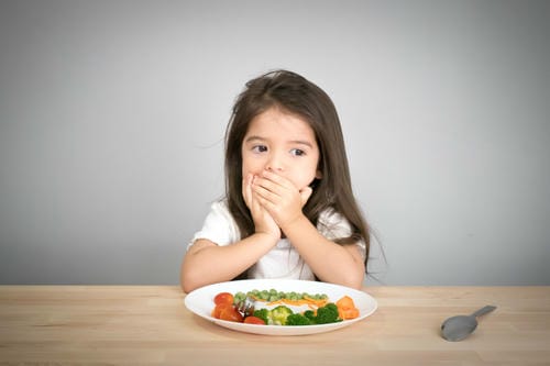 The girl presses both hands over her mouth, refusing to eat the mixed veggies (peas, broccoli, carrot and cherry tomatoes) laid on the plate in front of her.