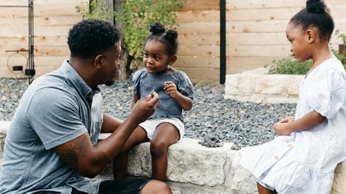 A father chats warmly with his two young daughters in a backyard garden. He holds a small object to draw the kids’ attention, creating a relaxed dialogue to guide and connect with the children.