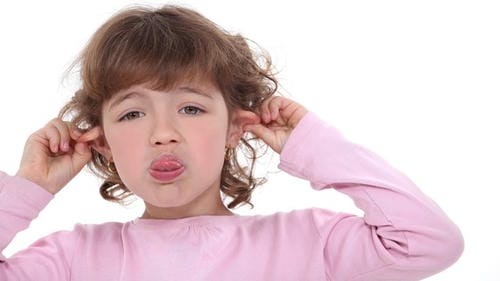 A curly-haired little girl pulls her ears with both hands and pouts her lips, making a playful, cheeky teasing face.