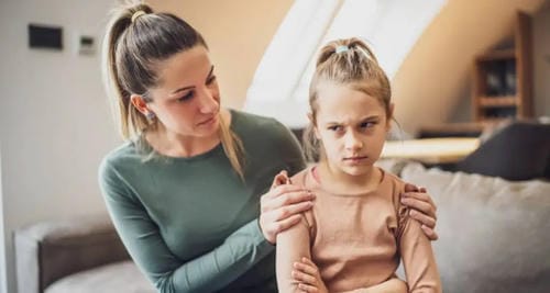 A mother gently holds her pre-teen daughter’s shoulders indoors to soothe her. The girl crosses her arms and wears a sulky, annoyed expression, showing she is upset or throwing a grumpy fit.