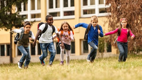 A diverse group of elementary students with backpacks run and leap cheerfully on the lawn outside their school building, full of excitement and energy after school hours.