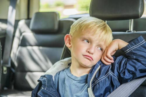 A young boy sits buckled in a car backseat, resting his head on one hand with a droopy, listless expression, feeling tired or bored on the ride home from school.