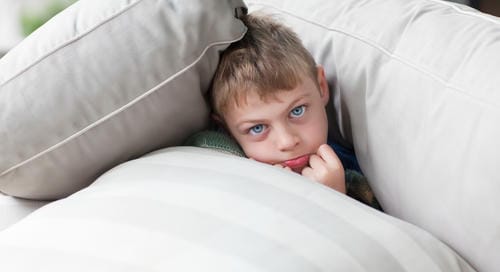 A young boy tucks himself between soft couch pillows, biting his finger and looking timid, showing shyness or mild anxiety.