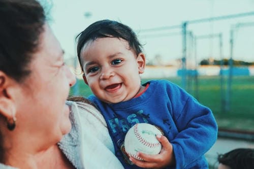 A joyful baby snuggles in an adult’s arms outdoors, gripping a baseball and smiling widely during warm parent-child interaction.