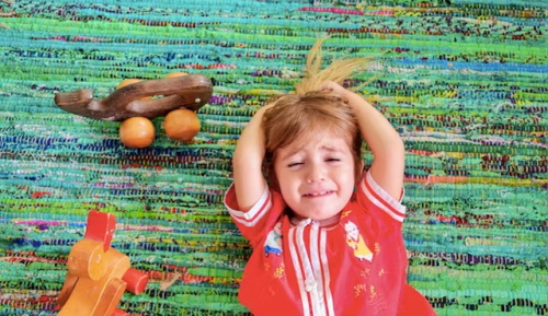 A young girl lies on a woven colorful rug beside wooden toy cars, clutching her hair and crying sadly, upset while playing alone indoors.