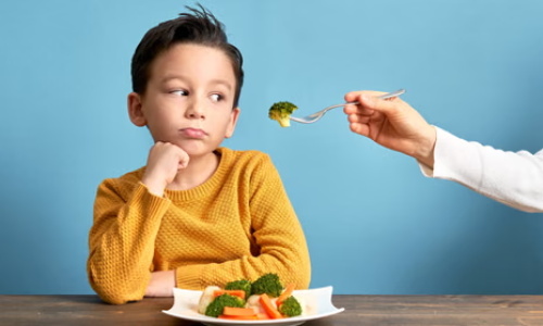 A young boy props his chin with his fist and turns his eyes away, showing obvious distaste as an adult holds a fork of broccoli toward his mouth; a plate of mixed cooked veggies sits in front of him.