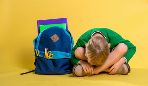 A young child curls up with knees drawn to the chest beside a full school backpack, worn out and emotionally drained from school pressure.
