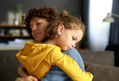 A mother hugs her gloomy daughter tightly on a sofa to soothe the kid’s negative feelings and ease her stress.